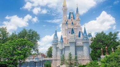 A fairy-tale castle with blue rooftops and golden spires stands surrounded by lush greenery and a moat under a bright blue sky with fluffy white clouds—perfect for capturing magical Disney Summer Tips.