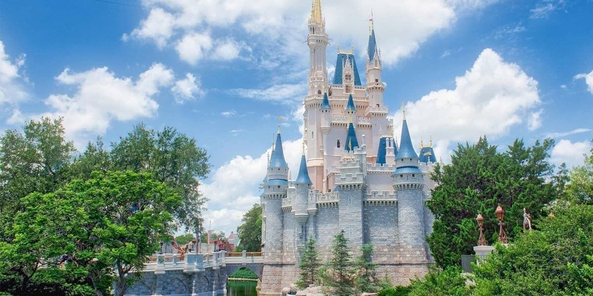 A fairy-tale castle with blue rooftops and golden spires stands surrounded by lush greenery and a moat under a bright blue sky with fluffy white clouds—perfect for capturing magical Disney Summer Tips.