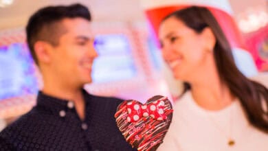 A man and woman smile at each other in the background, holding a heart-shaped chocolate treat with a red and white polka dot bow in the foreground—the perfect Disney Date night moment.