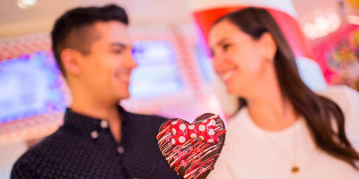 A man and woman smile at each other in the background, holding a heart-shaped chocolate treat with a red and white polka dot bow in the foreground—the perfect Disney Date night moment.