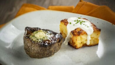 A plated meal reminiscent of the elegant offerings at underrated Disney restaurants: grilled filet steak with herb butter beside golden potato gratin in creamy sauce, garnished with chives, served on a white plate with an orange napkin in the background.