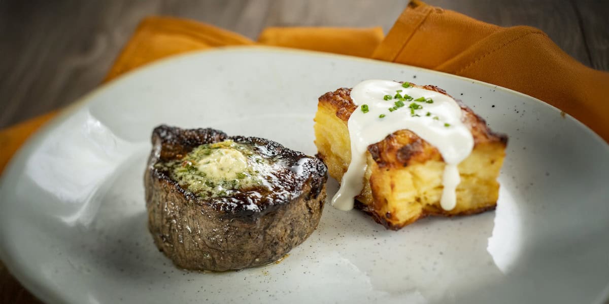 A plated meal reminiscent of the elegant offerings at underrated Disney restaurants: grilled filet steak with herb butter beside golden potato gratin in creamy sauce, garnished with chives, served on a white plate with an orange napkin in the background.