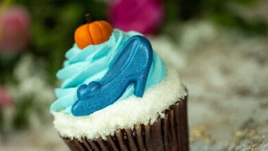 A cupcake topped with blue frosting, a small orange pumpkin decoration, and a blue candy high-heeled shoe—this whimsical treat could easily be among the Best Cupcakes at Disney World. The background is blurred with green and pink hues.