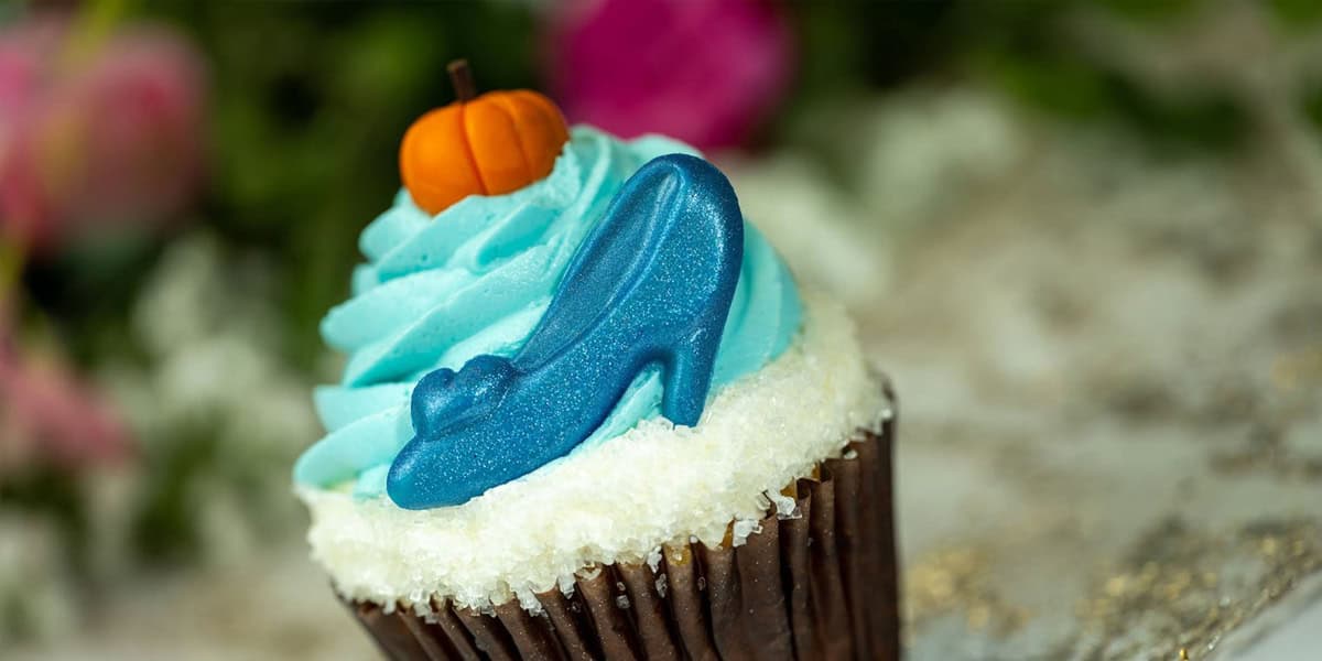 A cupcake topped with blue frosting, a small orange pumpkin decoration, and a blue candy high-heeled shoe—this whimsical treat could easily be among the Best Cupcakes at Disney World. The background is blurred with green and pink hues.
