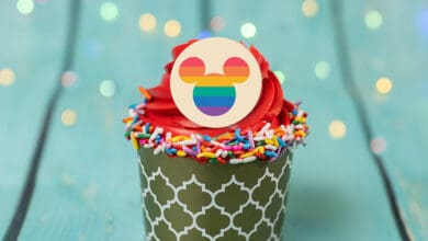 A cupcake with red frosting, rainbow sprinkles, and a round topper featuring a Mickey Mouse head with rainbow stripes, set against a teal wooden background—one of the Best Cupcakes at Disney World, glowing under soft, colorful lights.