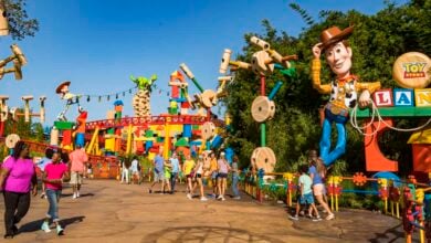 People walking along a path in Toy Story Land with colorful decorations. Large Woody figure holding a lasso seen on the right, Rex the dinosaur and other toys visible. Bright, sunny day with clear sky.