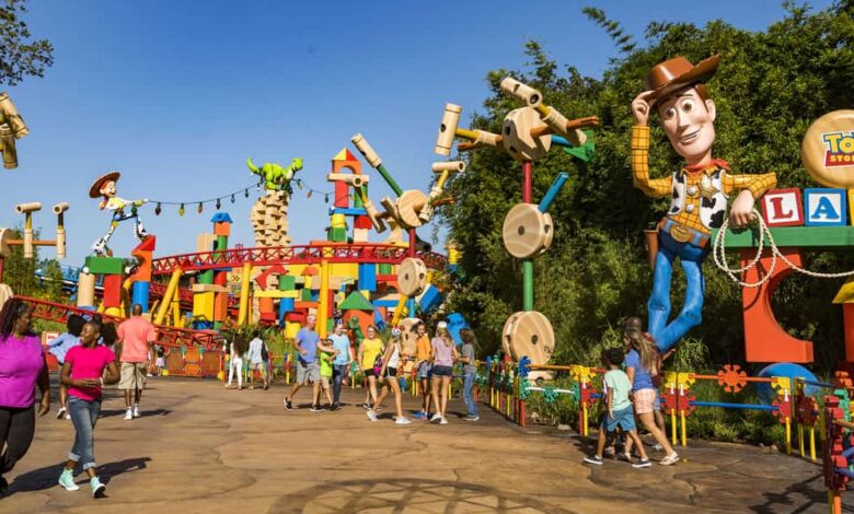 People walking along a path in Toy Story Land with colorful decorations. Large Woody figure holding a lasso seen on the right, Rex the dinosaur and other toys visible. Bright, sunny day with clear sky.
