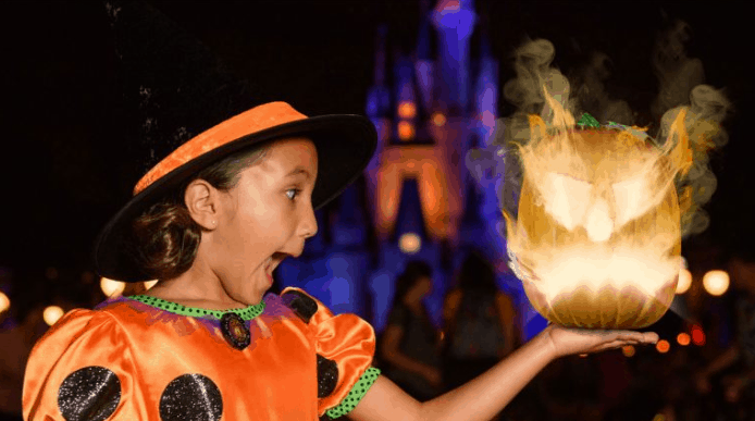 A young girl in a pumpkin-themed witch costume holds out her hand, looking surprised at a glowing, fiery jack-o'-lantern floating above it, with a brightly lit castle in the background at night.
