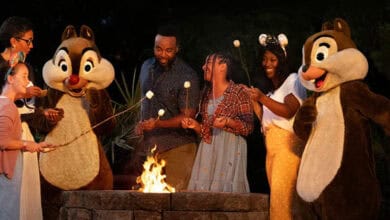 A group of people, including two children and two adults, roast marshmallows over a campfire with two people dressed as Chip and Dale characters, smiling and sharing Disney World Resort trivia while enjoying the evening outdoors.