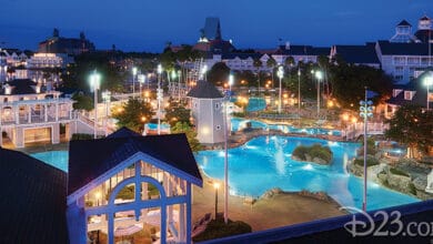 A large Disney World Resort shines at night, brightly lit with pools, waterfalls, lounge chairs, and peaked-roof buildings. The tranquil scene captures resort magic—perfect for Disney World Resort Trivia fans. Marked with a D23.com watermark.