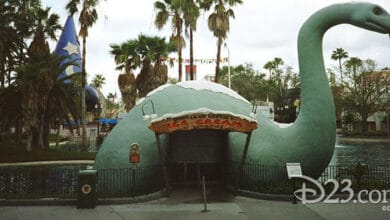 A large dinosaur-shaped building by a pond serves as an ice cream stand, with palm trees, a Sorcerer’s Hat structure, and colorful flags in the background—one of the Hollywood Studios secrets captured. The image is watermarked "D23.com" and "© Disney.