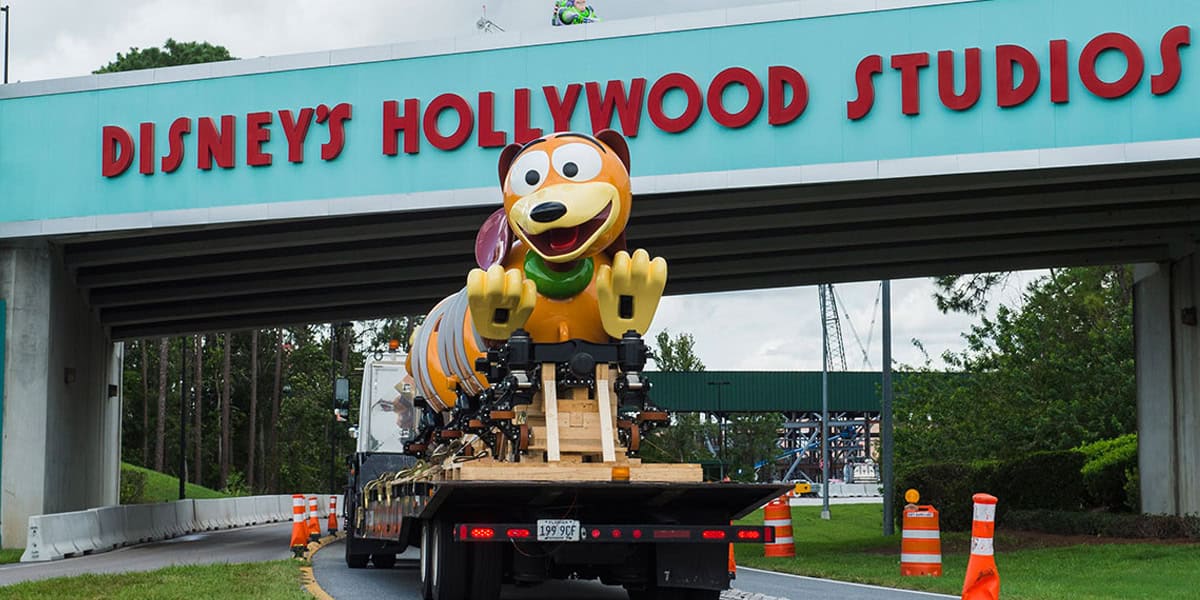 A large Slinky Dog ride vehicle from Toy Story is transported on a truck under the Disney's Hollywood Studios entrance sign, offering a glimpse into Hollywood Studios secrets amid trees and orange traffic cones.