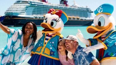 A smiling family takes a selfie with people in Daisy Duck and Donald Duck costumes on a sunny beach, with a large cruise ship in the background—perfect for capturing memories and gathering Disney Cruise Tips for your next adventure, as a new details emerge about a Disney cruise incident.