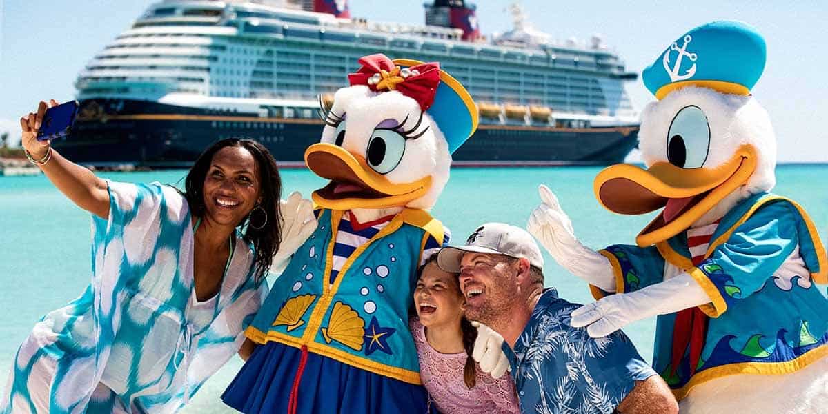 A family takes a selfie with people dressed as Disney characters Daisy Duck and Donald Duck on a sunny beach, with clear turquoise water and a Disney cruise ship in the background—capturing memories to cherish and gather Disney Cruise tips for their next voyage.