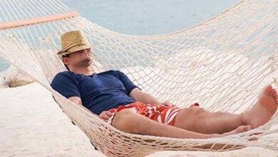A person wearing a straw hat, blue shirt, and red striped shorts is lying in a white hammock on a sandy beach, relaxing with eyes covered by the hat—an ideal scene for unwinding after exploring Disney Cruise Tips.