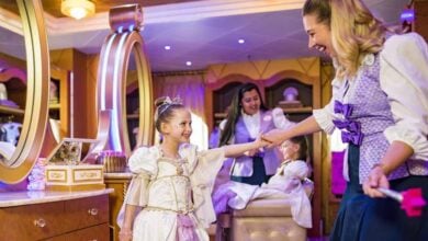 A young girl dressed as a princess smiles and holds hands with a woman in a salon setting, capturing the joy often seen at Disney Cruise Tips-inspired makeovers, while another girl gets her hair done by staff in the background.