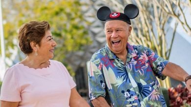 An older couple, proud Annual Passholders, smiles and enjoys a sunny day outdoors. The man wears a colorful Mickey Mouse shirt and Mickey ear hat, while the woman in a light pink shirt looks at him among trees and plants in the background.