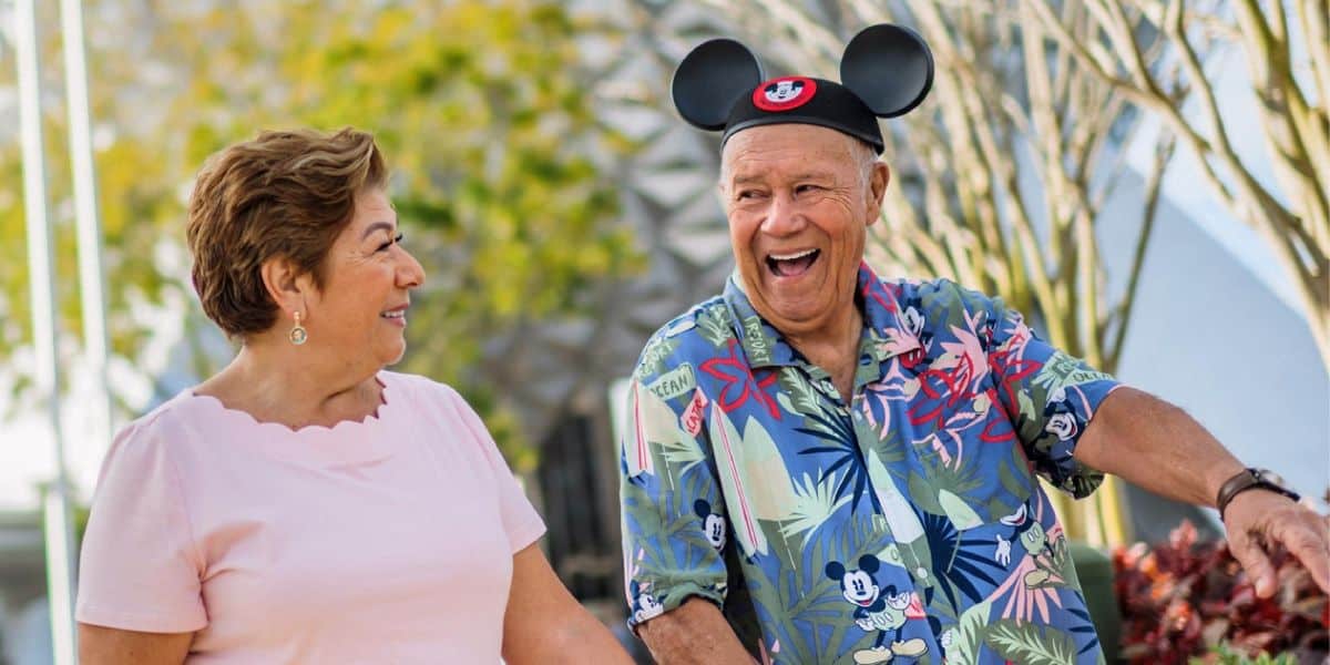 An older couple, proud Annual Passholders, smiles and enjoys a sunny day outdoors. The man wears a colorful Mickey Mouse shirt and Mickey ear hat, while the woman in a light pink shirt looks at him among trees and plants in the background.