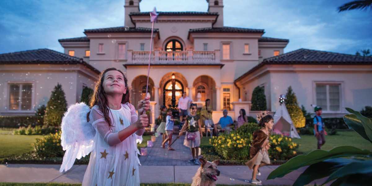 A young girl dressed as an angel, wearing wings and a starry costume, gazes upwards while holding a wand. She stands in front of a large, well-lit house at dusk. Several other children in costumes and a dog are seen playing around the yard.