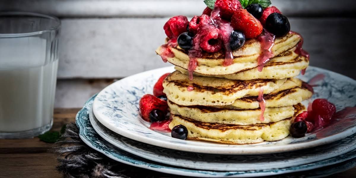 A stack of fluffy pancakes topped with fresh raspberries, strawberries, and blueberries, drizzled with berry sauce, sits on a decorative plate next to a glass of milk—a perfect Disney Springs Breakfast delight.