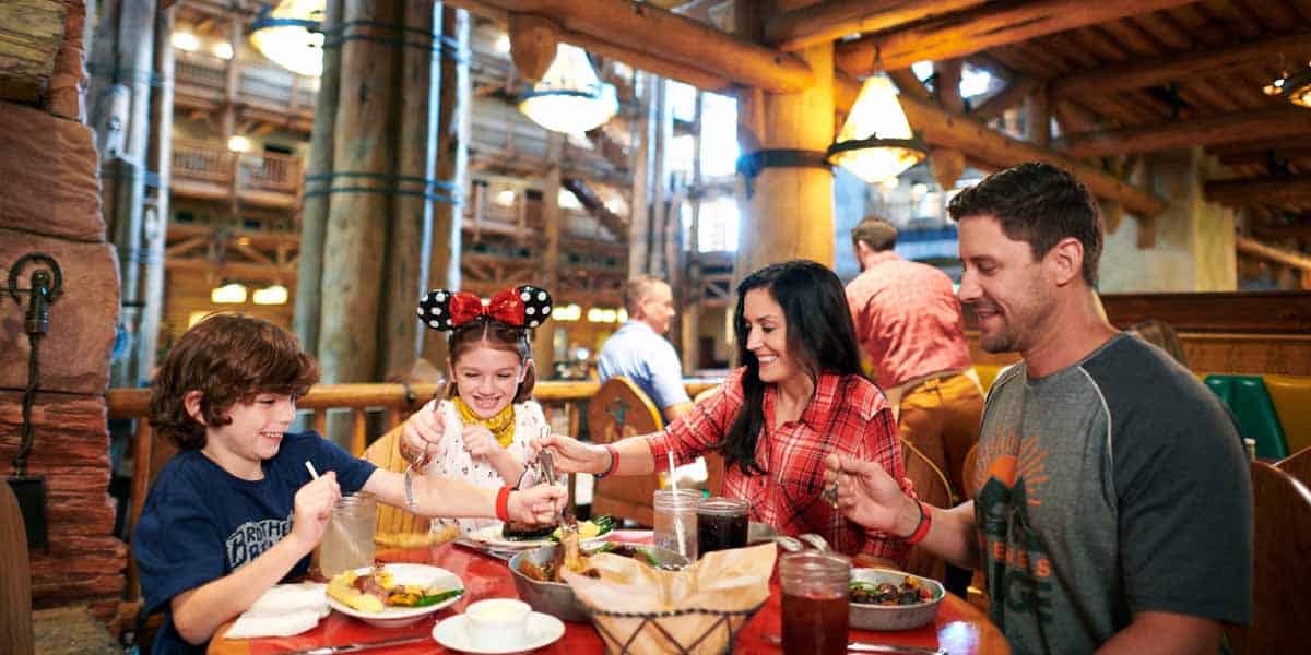 A family of four, including two children, sits at a rustic, lodge-style restaurant table, smiling and enjoying a meal together at a Disney World Table Service dining location.