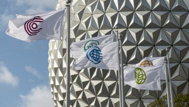 Four white flags with different colored circular symbols fly on flagpoles in front of a geometric, silver domed structure under a partly cloudy sky.