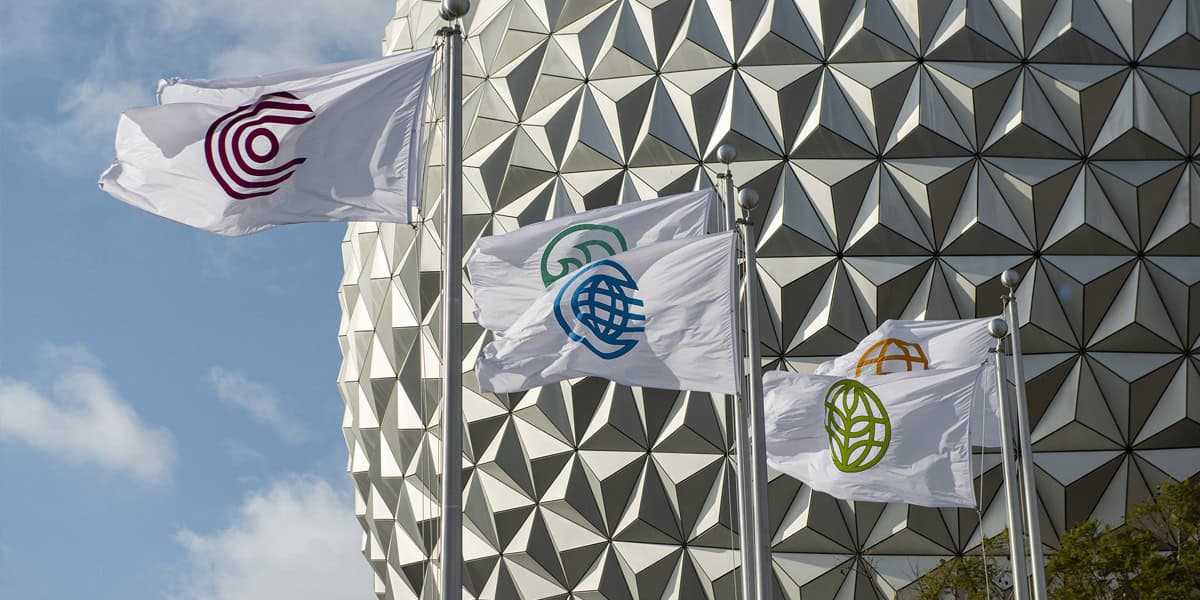 Four white flags with different colored circular symbols fly on flagpoles in front of a geometric, silver domed structure under a partly cloudy sky.