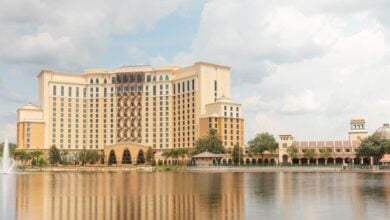 A large, elegant hotel with a beige and brown facade stands beside a calm lake. A fountain sprays water in the lake, and a smaller building with a domed roof is nearby. The sky is partly cloudy, enhancing the serene atmosphere.