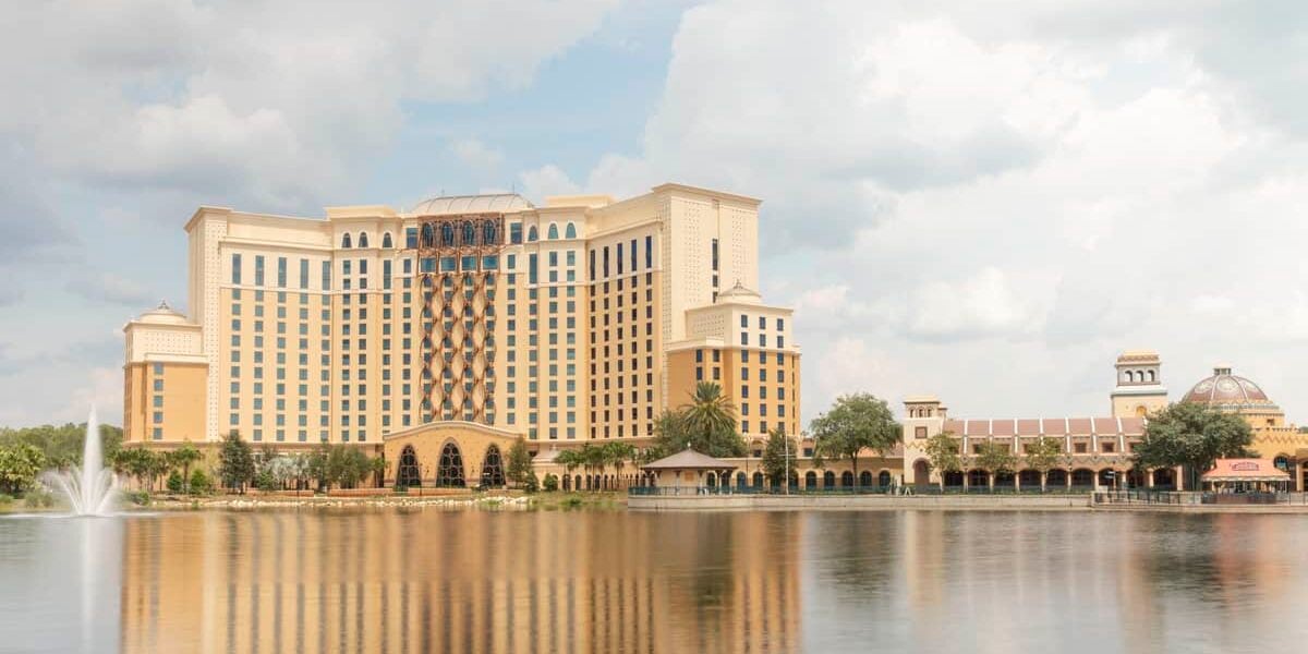 A large, elegant hotel with a beige and brown facade stands beside a calm lake. A fountain sprays water in the lake, and a smaller building with a domed roof is nearby. The sky is partly cloudy, enhancing the serene atmosphere for Disney guests.