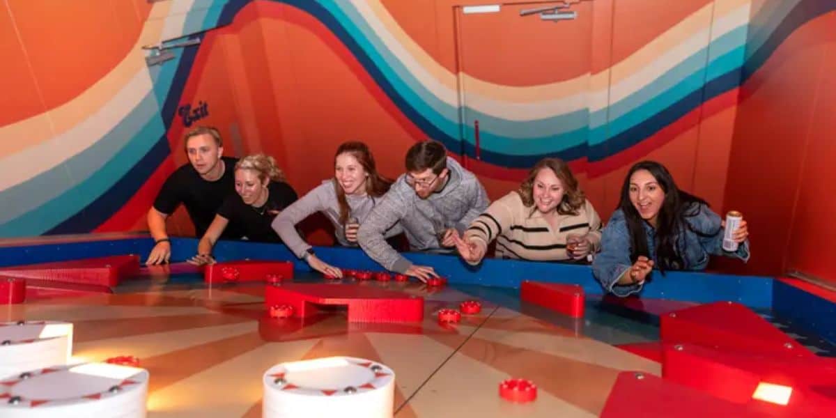 A group of six people joyfully playing an arcade game with red blocks on a brightly colored game surface. Some are holding drinks, and all are smiling or laughing, surrounded by vibrant orange and blue striped walls.