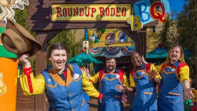 Four cheerful staff in colorful cowboy-themed uniforms pose and smile in front of the Roundup Rodeo BBQ restaurant entrance, which has playful Western and barbecue decorations—a picture-perfect spot for fans seeking fun Walt Disney World facts.
