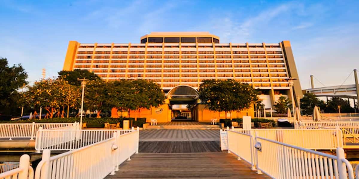 A large, modern hotel with a unique A-frame structure—Disney’s Contemporary Resort—is seen at sunset. The building is surrounded by trees and a marina, with a wooden walkway and white railings leading up to the entrance.