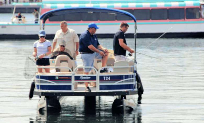 A group of four friends on a pontoon boat enjoys a serene fishing trip, casting their lines under the sun. It's not Walt Disney World, but this "guy’s trip" promises adventure, with one holding a net and another boat drifting peacefully in the background.