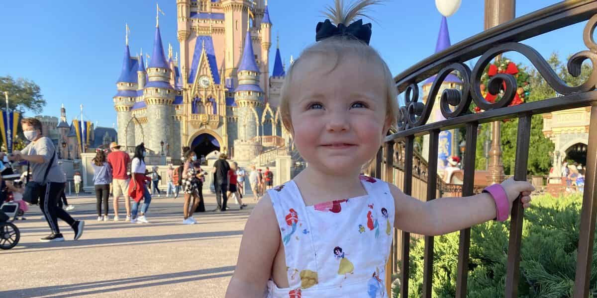 A smiling toddler wearing a dress with Disney princesses stands near a fence in front of Cinderella Castle at a Disney theme park, with people walking in the background.