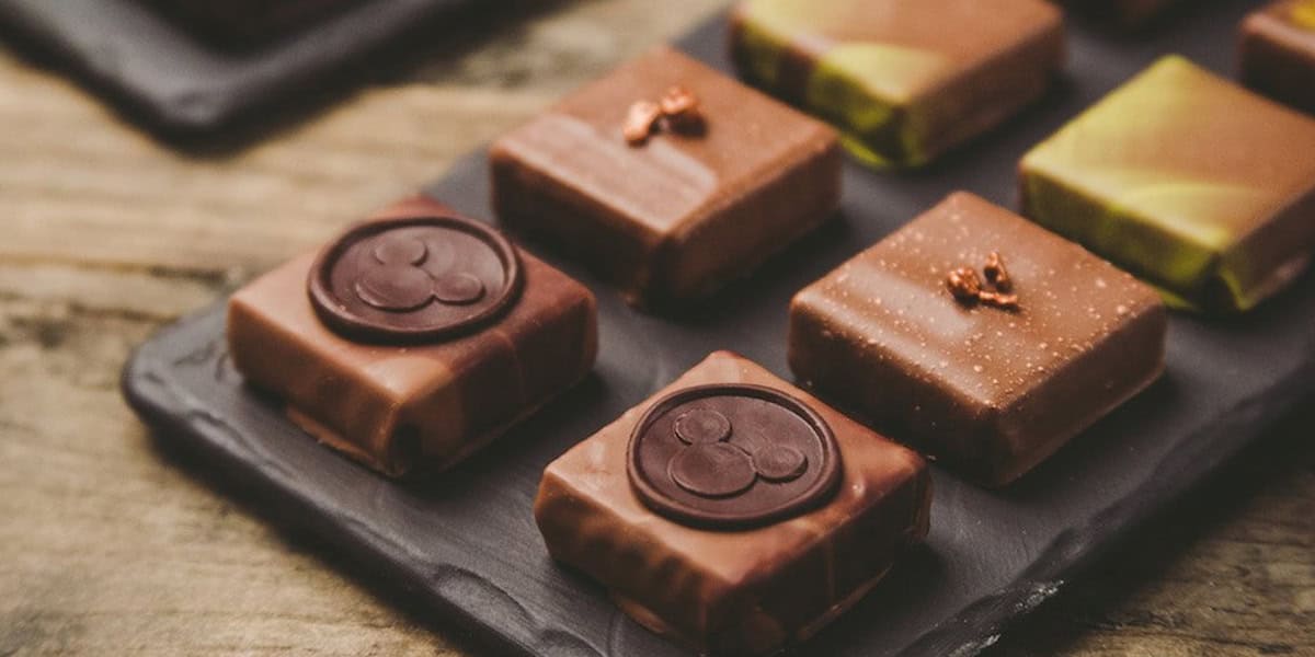 A close-up of assorted square chocolate treats at Disney Springs, arranged on a dark slate serving tray, featuring different toppings and decorative designs on a wooden table.