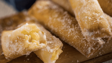 Close-up of sugar-coated fried pastry rolls on a wooden board, with one roll broken open to reveal a creamy filling inside.