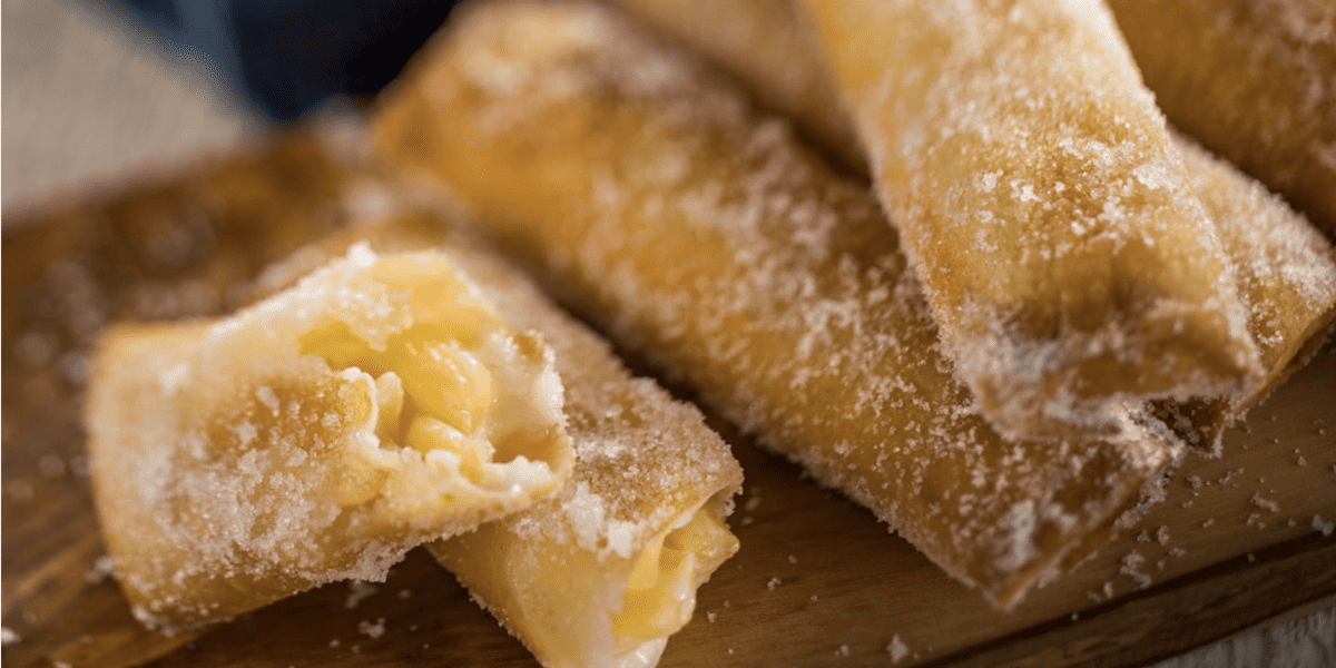 Close-up of sugar-coated fried pastry rolls on a wooden board, with one roll broken open to reveal a creamy filling inside.
