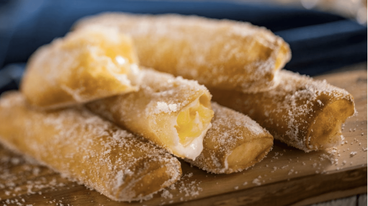 Four golden, sugar-dusted pastry rolls filled with creamy custard are stacked on a wooden surface. One pastry is split open, giving a glimpse of the rich filling—just like the treats in the Disney Pineapple Lumpia Recipe.