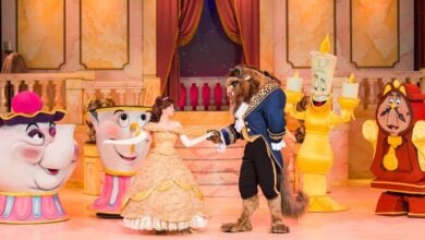 Characters from "Beauty and the Beast" perform on stage in a celebration of love. Belle, in a yellow gown, dances with the Beast. Mrs. Potts, Chip, Lumière, and Cogsworth are in the background. The set resembles Walt Disney World attractions with a grand ballroom featuring a red curtain and staircase.