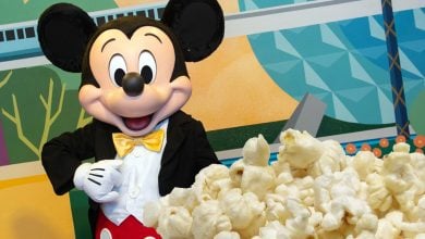 Mickey Mouse in a tuxedo with a yellow bow tie stands next to a large pile of popcorn, with a colorful geometric mural and hints of iconic Disney scents in the background.