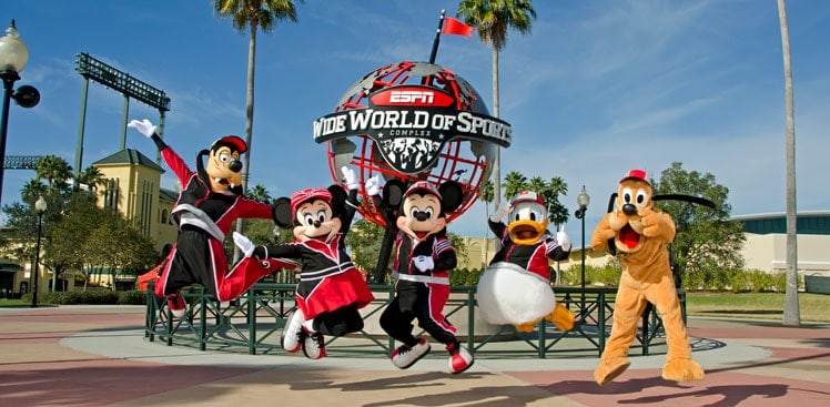 Mickey Mouse, Minnie Mouse, Donald Duck, Goofy, and Pluto joyfully leap in front of the ESPN Wide World of Sports globe structure under a clear sky and swaying palm trees, blissfully unaware of any Disney Wide World of Sports lawsuit.