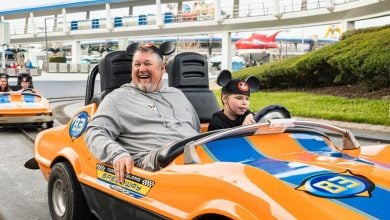 A man and a child wearing Mickey Mouse ears drive an orange and blue race car at an amusement park, enjoying one of the classic non-thrill Disney rides, with other people in similar cars following behind on the track.