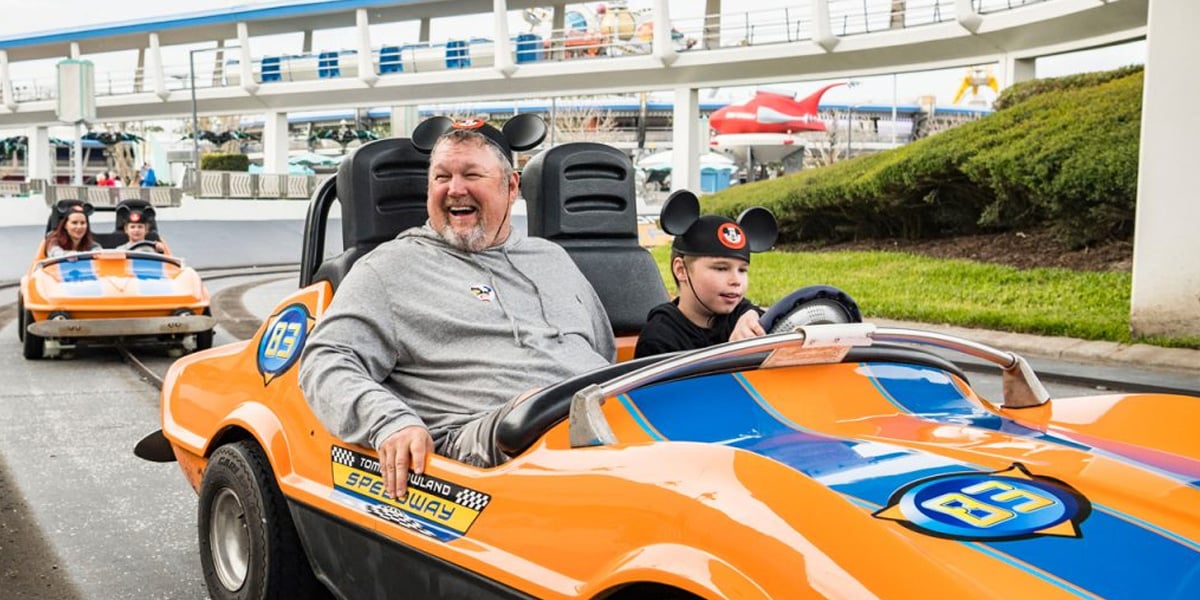 A man and a child wearing Mickey Mouse ears drive an orange and blue race car at an amusement park, enjoying one of the classic non-thrill Disney rides, with other people in similar cars following behind on the track.