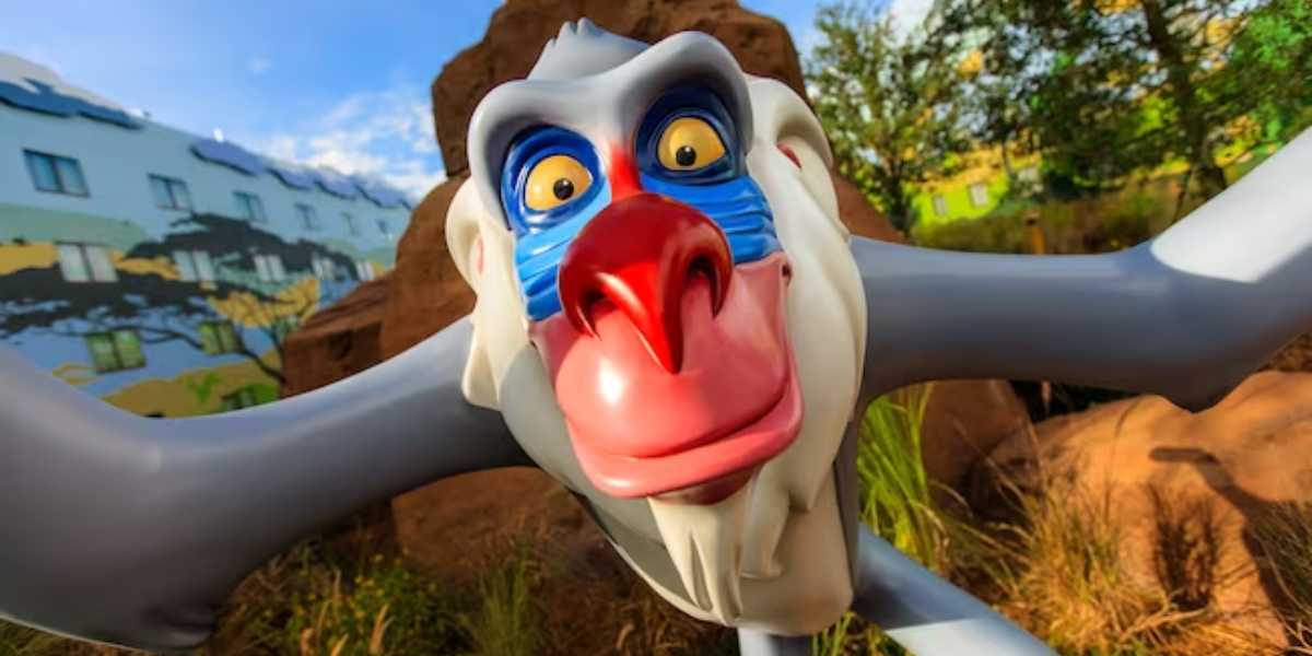 Close-up of a colorful statue of Rafiki, a baboon from Disney's The Lion King, at one of the Disney World Resorts. His blue, red, and white face is highlighted with an open, friendly expression and wide eyes. He's part of a themed area with lush greenery and rock structures in the background.