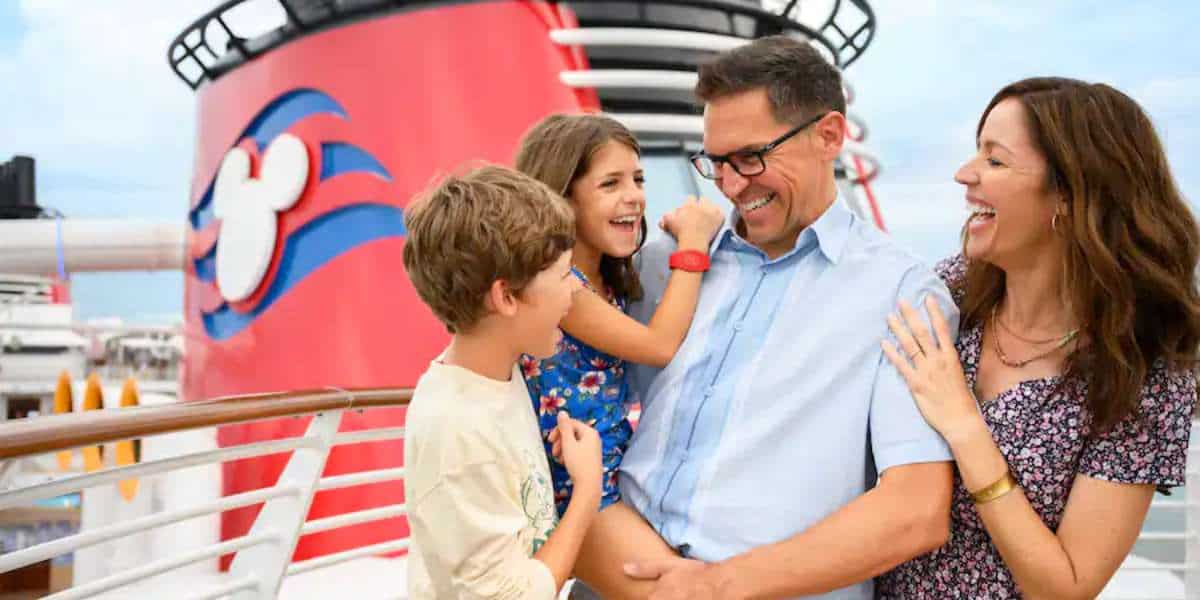 a brunette family of a small boy in a whte shirt, a long haired little girl in blue held by her dad in a blue button up and glasses and the mom smile aboard a disney cruise ship