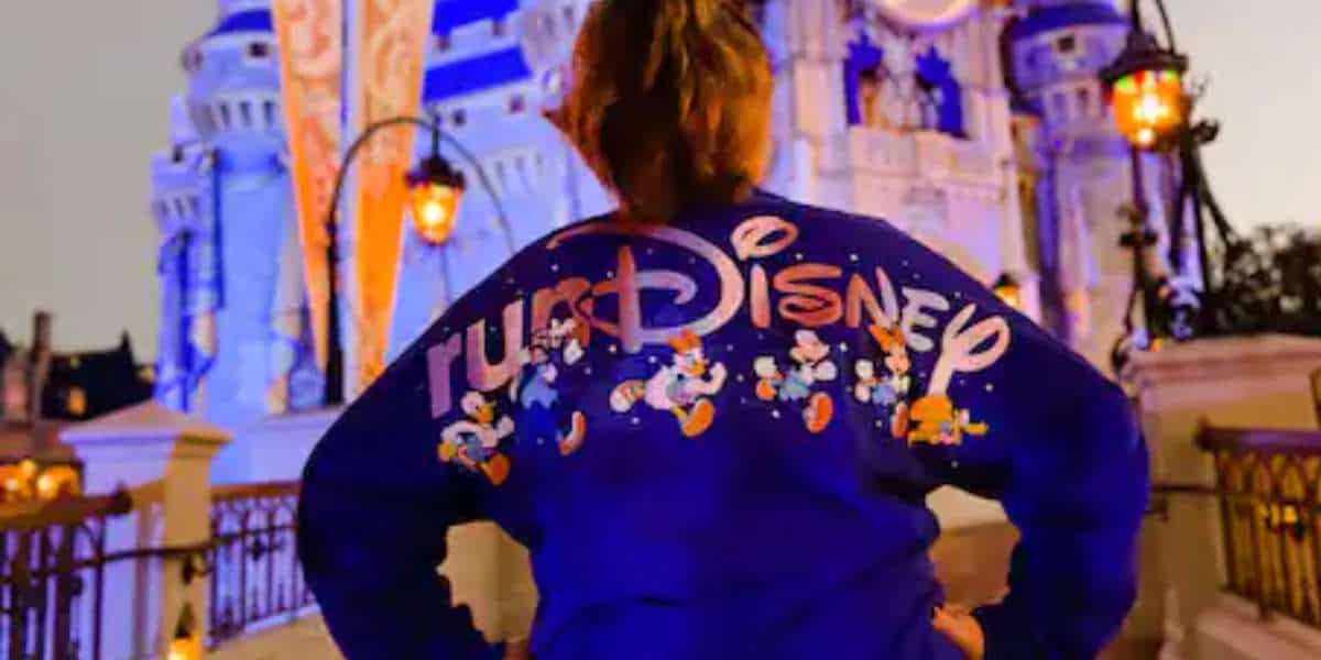 brown haired woman faces cinderella castle wearing a blue spirit hersey that reads RUNDISNEY with the fab five