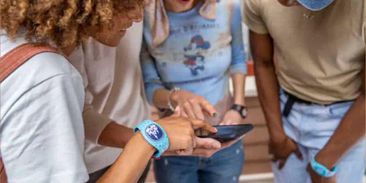 a group of four people around a mobile phone point at the screen, from left to right a woman with brown skin and curly blonde hair, another man in white, a white blonde woman with a blue long sleeve disney shirt, and a black man in a brown shirt and a blue magic band with jeans