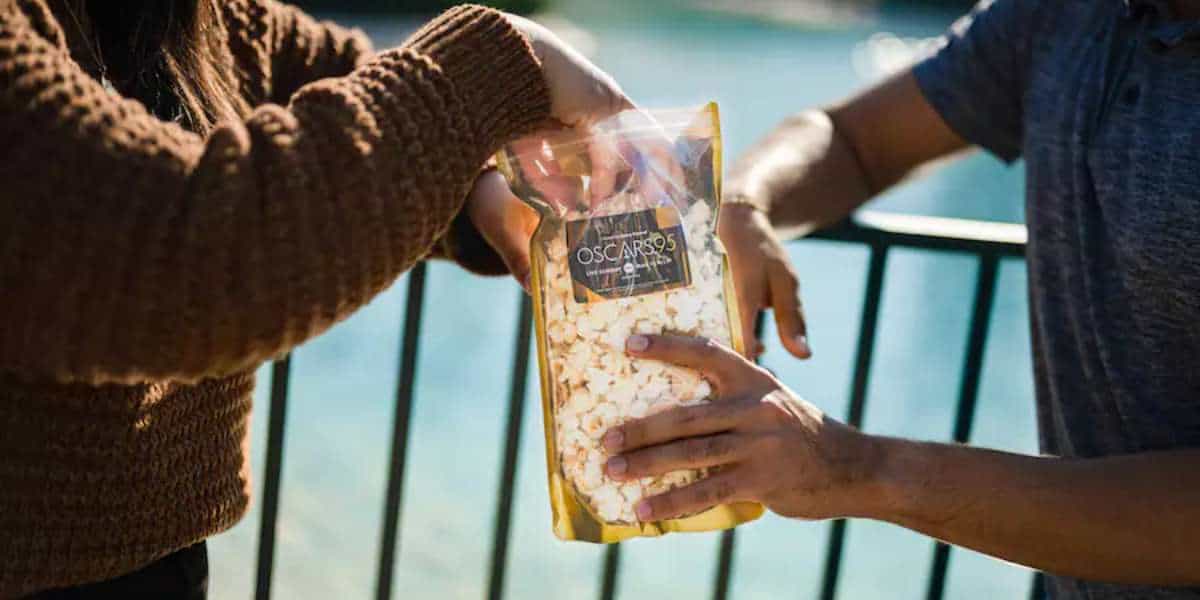 who hands enjoy a bag of popcorn with an oscars logo in front of a black fence and blue water