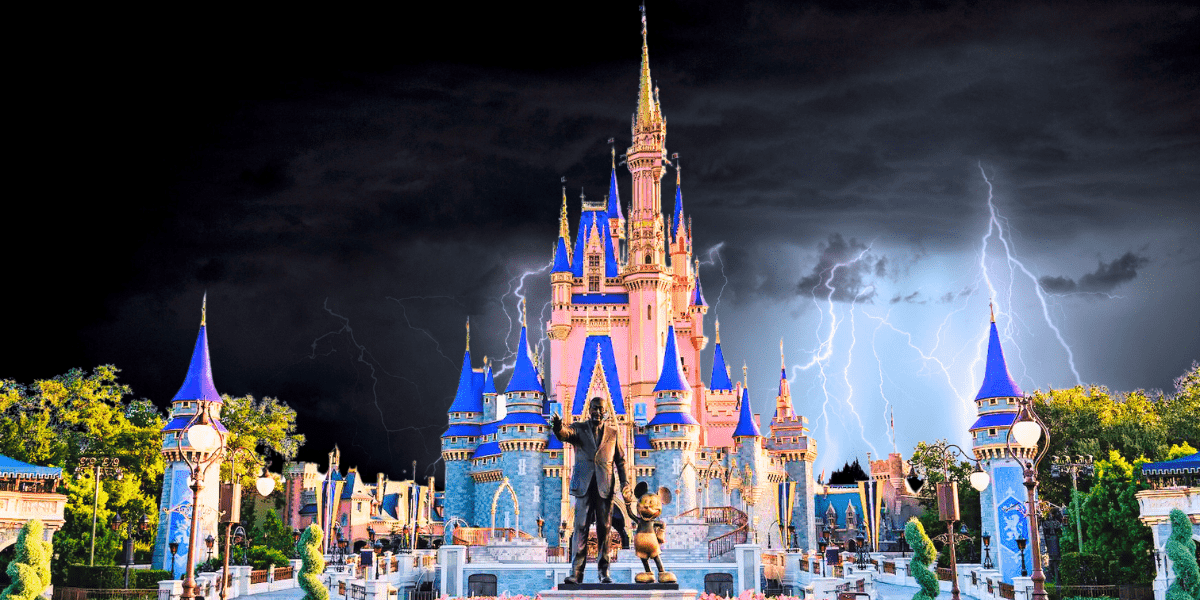 A brightly lit castle with blue and gold accents is the focal point under a dramatic dark sky. Lightning bolts flash behind the castle, creating a stark contrast with the cheerful, colorful surroundings. A statue stands in front of the castle, capturing the essence of Disney magic.