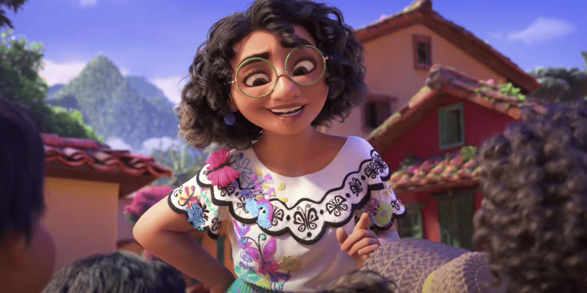 An animated character named Mirabel with curly hair and large round glasses smiles warmly. She wears a white embroidered blouse with colorful patterns and a pink tassel. In the background, there are reddish-orange buildings with tile roofs and lush green mountains and trees.